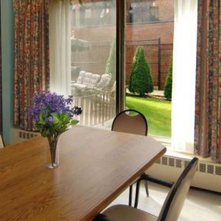 A cozy dining room with a wooden table, chairs, and a vase of flowers, overlooking a garden through large windows.