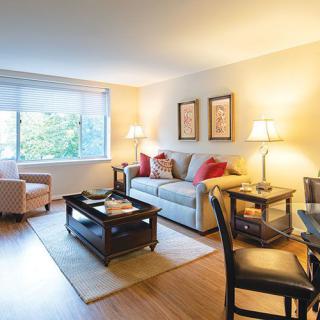 A cozy living room with a sofa, armchair, and coffee table, softly lit by lamps near a large window.