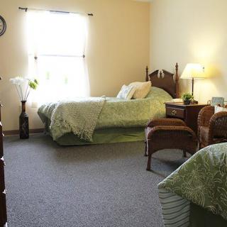 A cozy bedroom featuring twin beds with matching green bedding, illuminated by a table lamp and natural light from the window.