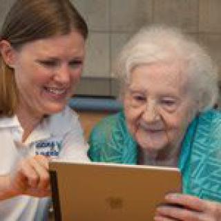 A young woman assists an elderly person in using a tablet indoors.