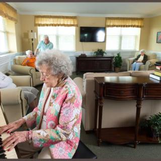 An elderly woman plays the piano in a cozy communal area of a retirement home, while other residents are seated and socializing.
