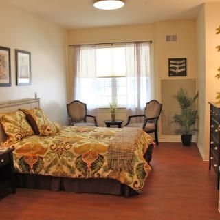 A cozy bedroom with elegant decor and wooden flooring, featuring a bed with patterned bedding, two chairs by the window, and framed artwork on the walls.