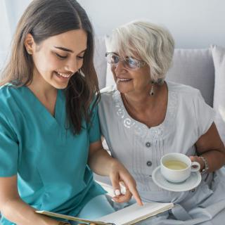 A nurse shares a light moment reading with an elderly woman enjoying a cup of tea.