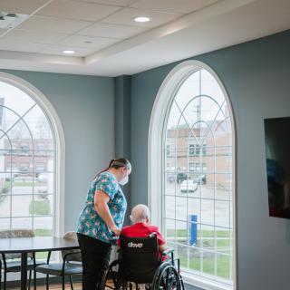 A caregiver assists an elderly person in a wheelchair by the window of a nursing home.