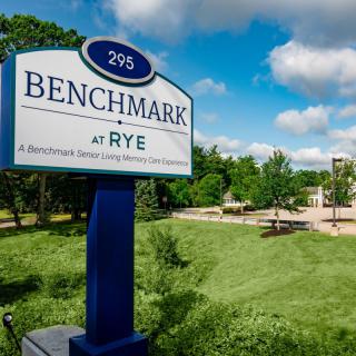 A sign for Benchmark at Rye is displayed near a grassy area with trees and a building in the background.