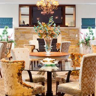 An elegant dining room featuring a round glass table surrounded by ornate chairs, adorned with a floral centerpiece and illuminated by a chandelier.