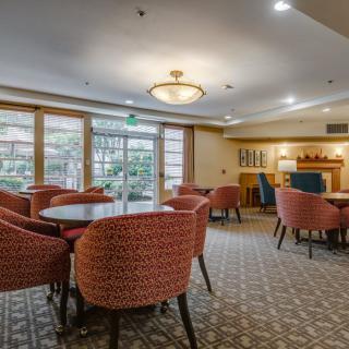 A well-lit dining area with round tables and upholstered chairs, featuring large windows and decorative lighting.