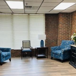 A cozy waiting area featuring blue patterned chairs, a small side table with a lamp, and a brick wall backdrop.