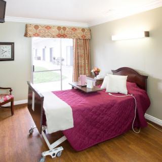 A well-lit hospital room featuring a bed with a burgundy cover, a chair, and large windows.