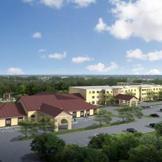 An aerial view of a large building complex with a parking lot, surrounded by trees under a partly cloudy sky.