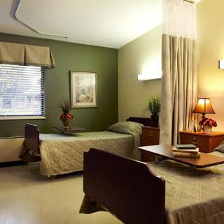 A cozy hospital room with two beds and green walls, featuring a lamp and a table.