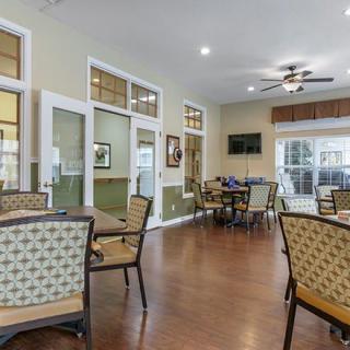 A cozy dining area with tables, chairs, and a ceiling fan, featuring large windows and a television.