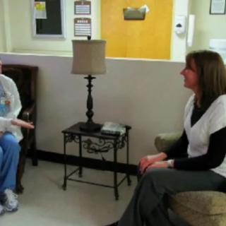 Two individuals are having a discussion in an office setting, sitting on chairs with a small table and lamp between them.
