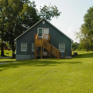 A small house with exterior wooden stairs surrounded by a lush green lawn and trees.