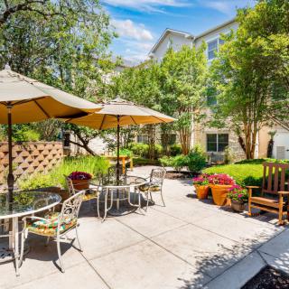 A sunny garden patio with tables and chairs under umbrellas, surrounded by lush greenery.