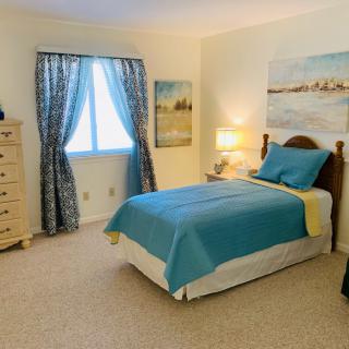 A cozy bedroom with a single bed featuring blue bedding, a dresser, and a warmly lit lamp on the nightstand.