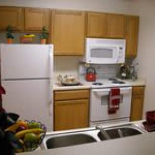 A cozy kitchen featuring wooden cabinets, a refrigerator, microwave, and oven, with a sink in the foreground and fruit displayed in a basket.