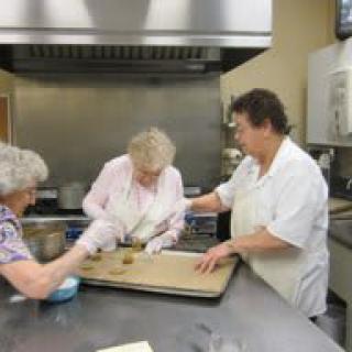 Three elderly women work together in a kitchen to prepare baked goods.