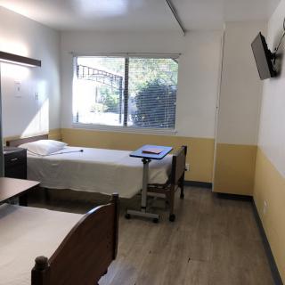 A simple hospital room with a single bed, a window, and a wall-mounted television.