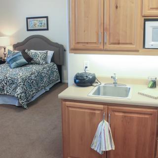 A cozy bedroom with a small kitchenette, featuring a bed, lamp, and sink.