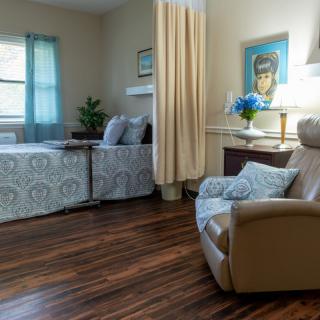 A cozy hospital room features two beds with a curtain divider, an armchair, and warm decor.