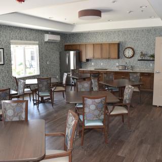 A cozy dining room with wooden tables and chairs, featuring a kitchenette and a wall clock.