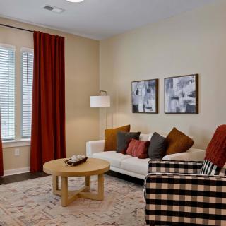 A cozy living room with a sofa, coffee table, armchair, and red curtains, accented by throw pillows and artwork on the wall.