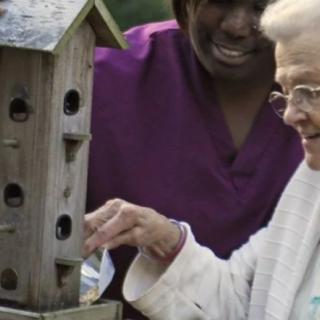 An elderly person and a caregiver interacting with a multi-level birdhouse outdoors.