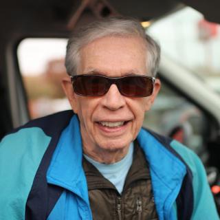 A man wearing sunglasses and a jacket smiles outdoors.