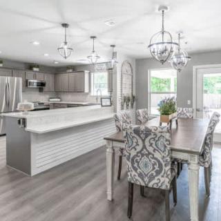 A modern open-concept kitchen and dining area with sleek gray tones, featuring chandeliers, stainless steel appliances, and elegant patterned chairs.