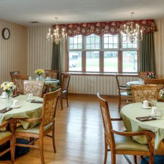 A cozy dining room with elegantly set tables and chairs, featuring chandeliers and large windows draped with curtains.