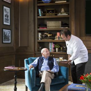 An elderly man sitting in an armchair in a library is being served by a waiter.