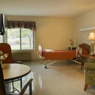 A well-lit hospital room with a bed, table, chairs, and a lamp by the window.