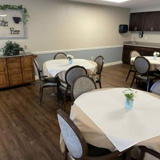 A cozy dining room with neatly arranged tables and chairs, featuring wooden flooring and a small kitchen area in the background.