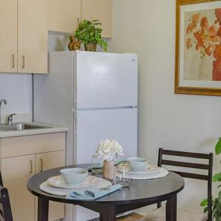 A cozy kitchen with a dining table set for two, featuring a refrigerator, plants, and wall artwork.