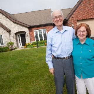 An elderly couple smiles happily outside their home.