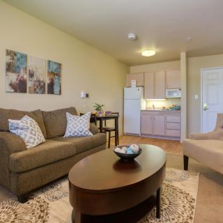 A cozy living room with a sofa, oval coffee table, and a glimpse of the kitchenette in a compact apartment.