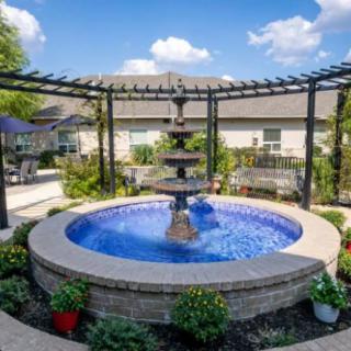 A serene garden setting featuring a central fountain surrounded by a pergola and lush greenery.