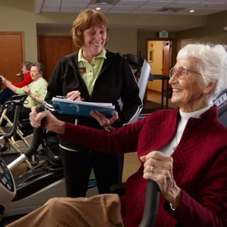 An elderly woman exercises on a stationary bike with the guidance of an instructor in a gym setting.
