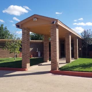 A well-structured brick building with a covered entrance, surrounded by green grass and under a clear blue sky.