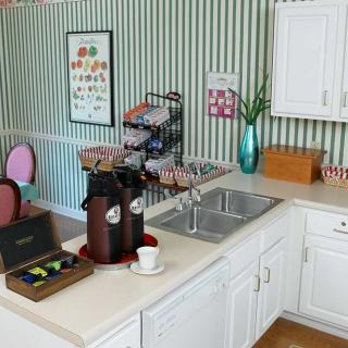 A cozy kitchen corner featuring a coffee station with dispensers, cups, and various condiments on a countertop next to a sink, all set against striped wallpaper and white cabinetry.