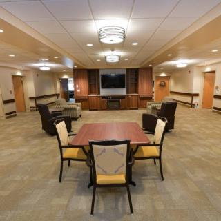 A spacious and well-lit lounge area featuring a central table surrounded by chairs, with a television and seating in the background.