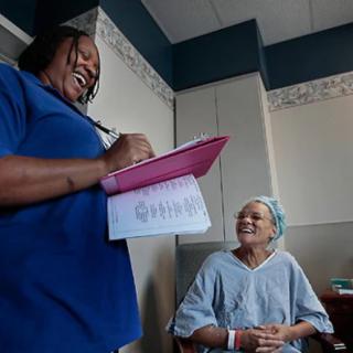 A nurse is interacting with a patient in a hospital room, holding a clipboard and smiling.