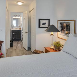A modern bedroom featuring a neatly made bed, bedside table with a lamp, and framed artwork on the wall. The view extends into a hallway leading to a bathroom.