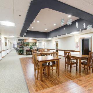 A spacious dining area with wooden tables and chairs, featuring a wood floor and ceiling lights, connected to a corridor.