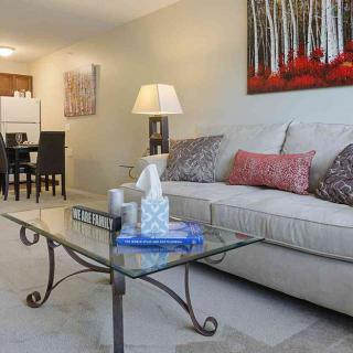 A cozy living room with a glass coffee table, comfortable sofa, and warm lighting leading to a dining area.