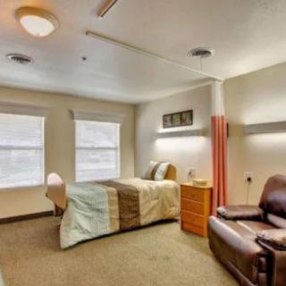 A cozy bedroom with a bed, chair, and natural light coming through two windows.