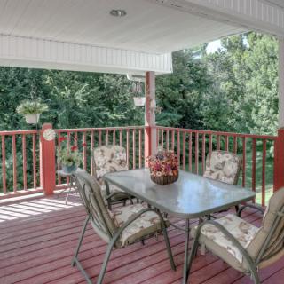 A cozy covered deck with a glass table and four cushioned chairs, surrounded by wooden railings and overlooking lush greenery.