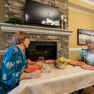 Two people sitting at a dining table having a conversation in a cozy room with a fireplace and television.