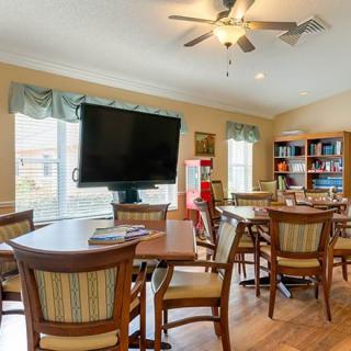 A cozy community room with multiple tables and chairs, a large television, bookshelves, and ample natural light from the windows.